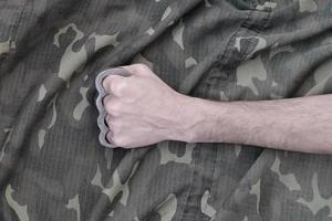 Male fist with brass knuckles on the background of a camouflage jacket. The concept of skinhead culture, handmade melee weapons photo