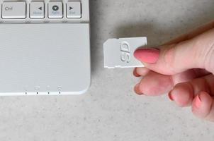 una mano femenina inserta una tarjeta sd compacta blanca en la entrada correspondiente en el lateral de la netbook blanca. mujer utiliza tecnologías modernas para almacenar memoria y datos digitales foto