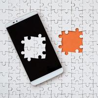 A modern big smartphone with several puzzle elements on the touch screen lies on a white jigsaw puzzle in an assembled state with missing elements photo