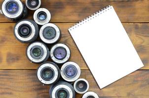 Several photographic lenses and white notebook lie on a brown wooden background. Space for text photo