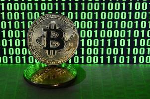 A pair of bitcoins lies on a cardboard surface on the background of a monitor depicting a binary code of bright green zeros and one units on a black background. Low key lighting photo