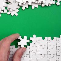 The hand folds a white jigsaw puzzle and a pile of uncombed puzzle pieces lies against the background of the green surface. Texture photo with space for text
