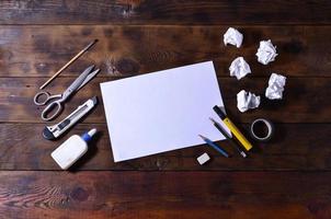 A school or office still life with a white blank sheet of paper and many office supplies. The school supplies lie on a brown wooden background. Place for text photo