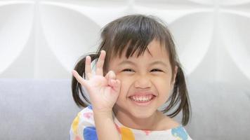 retrato de feliz y encantadora niña asiática de 4 años, pequeña niña preescolar con adorables coletas sonriendo mirando a la cámara mostrando los dedos bien con espacio para copiar. foto