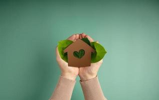 Eco House, Carbon Neutral Concepts. Clean Energy. Green Power. Hands Holding a Craft Papaer House and Green Leaf. Sustainable Resources, Environmental Care. Top View photo