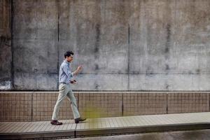 sonriente joven hombre de negocios asiático en ropa casual usando teléfono móvil mientras camina por la pared del edificio urbano. estilo de vida de la gente moderna. vista lateral. longitud total foto