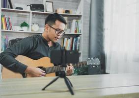 un profesor de música asiático transmite lecciones de guitarra en vivo a los estudiantes en línea usando el móvil. músico masculino asiático tocando guitarra cantando videoconferencias en vivo en casa. música de concierto en vivo desde cualquier lugar. foto