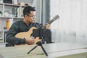 un profesor de música asiático transmite lecciones de guitarra en vivo a los estudiantes en línea usando el móvil. músico masculino asiático tocando guitarra cantando videoconferencias en vivo en casa. música de concierto en vivo desde cualquier lugar. foto