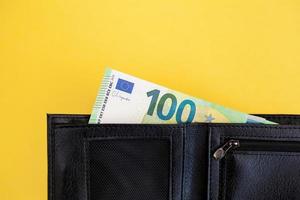 A hundred euro banknote sticks out of a black wallet on a yellow background. cash paper currency, payment, earning and savings, european currency, money and finance concept. space for text photo