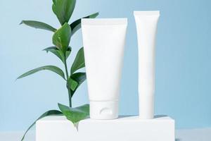 Plastic white tubes for face and eye cream or lotion. Skin care or sunscreen cosmetic on white podium on blue background with ruscus leaves. Beauty concept for face care photo