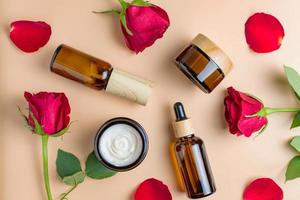 conjunto de productos de belleza de spa orgánicos naturales sobre fondo beige con rosas rojas y pétalos. aceite de rosa casero, tarro de crema hidratante, botella de spray de vidrio ámbar sobre fondo beige foto