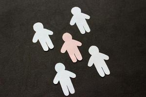 Silhouettes of five men, cut from paper. Between the white men there is one pink man. On black background photo