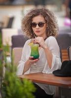 Happy Brunette Girl with sunglasses Sitting at the Park, Drinking a glass of Cold Green Juice While Smiling Into the Distance. Young pretty woman on the bench  drinking  juice wearing a white shirt photo