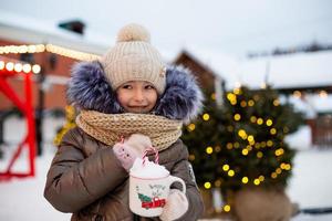 Girl with mug with snow, candy cane and inscription Merry and Bright in her hands outdoor in warm clothes in winter at festive market. Fairy lights garlands decorated snow town for new year. Christmas photo