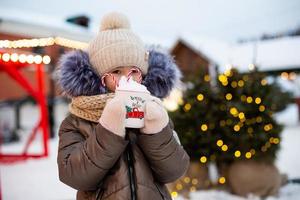 Girl with mug with snow, candy cane and inscription Merry and Bright in her hands outdoor in warm clothes in winter at festive market. Fairy lights garlands decorated snow town for new year. Christmas