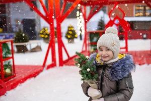 A girl with a Christmas tree in her hands outdoor in warm clothes in winter at a festive market. Fairy lights garlands decorated snow town for the new year photo