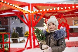 A girl with a Christmas tree in her hands outdoor in warm clothes in winter at a festive market. Fairy lights garlands decorated snow town for the new year photo