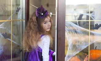 A girl in a witch costume on the windowsill by the window is playing with a spider web and a spider, decorating the house for the Halloween holiday. Terrible scenery, fear and horror, pumpkins photo