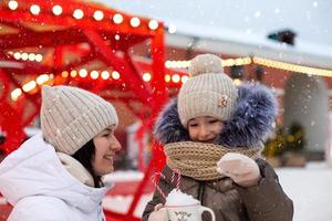 Mom and daughter for Christmas outdoor in warm clothes in winter at festive market. Fairy lights garlands decorated snow town for new year photo