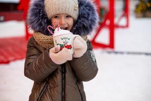 Girl with mug with snow, candy cane and inscription Merry and Bright in her hands outdoor in warm clothes in winter at festive market. Fairy lights garlands decorated snow town for new year. Christmas photo