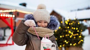 Girl with mug with snow, candy cane and inscription Merry and Bright in her hands outdoor in warm clothes in winter at festive market. Fairy lights garlands decorated snow town for new year. Christmas photo