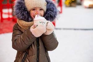 chica con taza con nieve, bastón de caramelo e inscripción alegre y brillante en sus manos al aire libre con ropa de abrigo en invierno en el mercado festivo. Guirnaldas de luces de hadas decoradas ciudad de nieve para año nuevo. Navidad foto