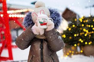 Girl with mug with snow, candy cane and inscription Merry and Bright in her hands outdoor in warm clothes in winter at festive market. Fairy lights garlands decorated snow town for new year. Christmas photo