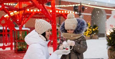 Mom and daughter for Christmas outdoor in warm clothes in winter at festive market. Fairy lights garlands decorated snow town for new year photo