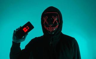 concepto de seguridad digital. hacker anónimo con máscara sosteniendo un teléfono inteligente pirateado. foto