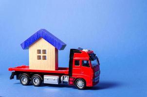 Red truck carrier transports a blue roofed house. Concept of transportation and cargo shipping, moving company. Construction of new houses and objects. Logistics and supply. Move entire buildings photo