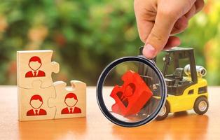 A magnifying glass looks at a forklift truck carries a red puzzle to the unfinished assembly of business team. Search, recruitment staff, hiring leader. Creating efficient and productive business unit photo