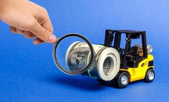 A magnifying glass looks at a forklift truck carries a bundle of dollars. Attracting direct investment in business and production, improving economic performance. capitalism. Export of capital photo