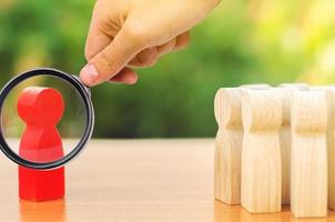 A magnifying glass looks at a red figurine of a man stands aside from the crowd of people. Asociality, sociopathy. Development of leadership and social qualities. Rejected from society, lonely. photo