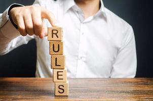 A man holds a tower of blocks with the word Rules from falling. Setting clear rule and restrictions. Leadership and discipline. Authoritarianism, tight control framework. Norms and laws in society photo