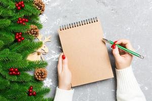 vista superior de la mano femenina escribiendo en un cuaderno sobre fondo de Navidad de cemento. abeto y decoraciones festivas. lista de deseos concepto de año nuevo foto