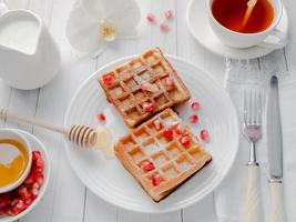 Delicious mouth-watering Viennese waffles with honey and pomegranate seeds on a white plate, light wooden background photo