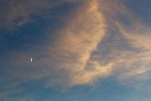 una luna creciente en el cielo azul entre las nubes iluminadas por el sol al atardecer foto