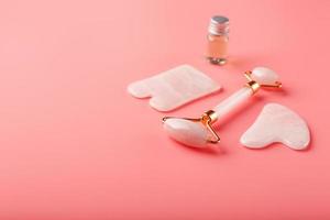 un conjunto de herramientas para la técnica de masaje facial gua sha hechas de cuarzo rosa natural sobre un fondo rosa. rodillo, piedra de jade y aceite en tarro de cristal para el cuidado facial y corporal.