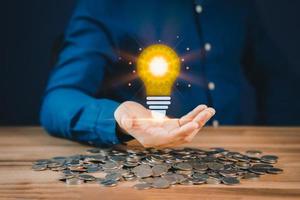 Man holding a glowing abstract light bulb with pile money cions on a wooden table, business problem solving ideas and creative marketing ideas Inventing and learning new things. photo