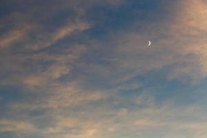una luna creciente en el cielo azul entre las nubes iluminadas por el sol al atardecer foto