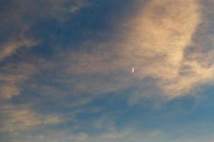 una luna creciente en el cielo azul entre las nubes iluminadas por el sol al atardecer foto