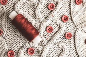Beautiful texture of a soft warm natural sweater, fabrics with a knitted pattern of yarn and red small round buttons for sewing and a skein of red thread. The background photo