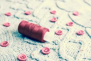 Beautiful texture of a soft warm natural sweater, fabrics with a knitted pattern of yarn and red small round buttons for sewing and a skein of red thread. The background photo