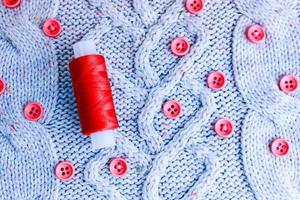 Beautiful texture of a soft warm natural sweater, fabrics with a knitted pattern of yarn and red small round buttons for sewing and a skein of red thread. The background photo