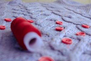 Beautiful texture of a soft warm natural sweater, fabrics with a knitted pattern of yarn and red small round buttons for sewing and a skein of red thread. The background photo