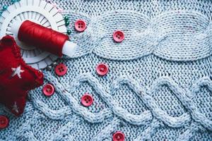 Beautiful texture of a soft warm natural sweater, fabrics with a knitted pattern and red small round buttons for sewing and a skein of thread, needle bed and needle pad. Flat lay. The background photo