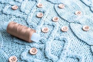 Beautiful texture of a soft warm natural sweater, fabrics with a knitted pattern of yarn and red small round buttons for sewing and a skein of red thread. The background photo