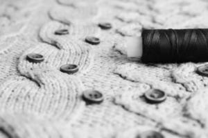 Beautiful black and white texture of a soft warm natural sweater, fabrics with a knitted pattern of threads and red small round buttons for sewing and a skein of thread. The background photo