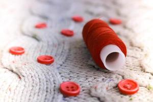 Beautiful texture of a soft warm natural sweater, fabrics with a knitted pattern of yarn and red small round buttons for sewing and a skein of red thread. The background photo