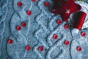 Beautiful texture of a soft warm natural sweater, fabrics with a knitted pattern and red small round buttons for sewing and a skein of thread, needle bed and needle pad. Flat lay. The background photo